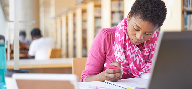 Aluna de curso de educação a distância está estudando em frente a tela de computador. Ela tem cabelos crespos e curtos e usa lenço rosa estampado no pescoço.