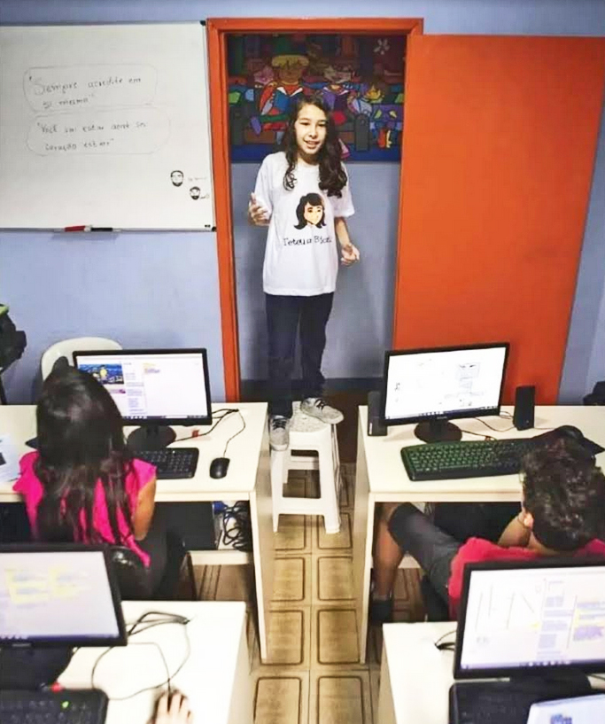 Imagem mostra Teteus Bionic em pé na frente dasala de aula, com crianças sentadas em mesas com computadores.