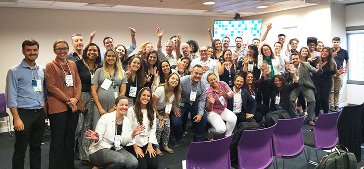 Grupo de 50 pessoas que participaram do evento Prata da Casa, que destacou ações pela inclusão do Programa de Voluntariado da Fundação Telefônica Vivo, estão sorrindo e acenando para a foto.