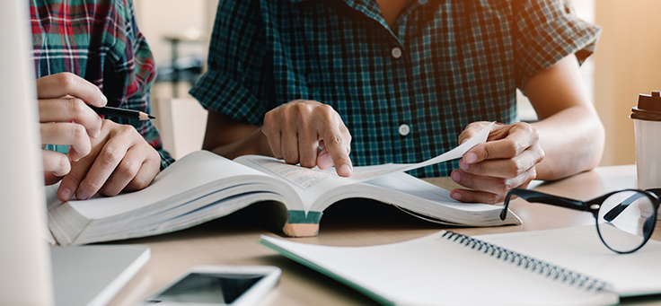 Na imagem duas pessoas estão sentadas lado a lado procurando informações em um livro e há um óculos e um caderno sobre a mesa. Conheça e ressignifique 11 termos sobre voluntariado.