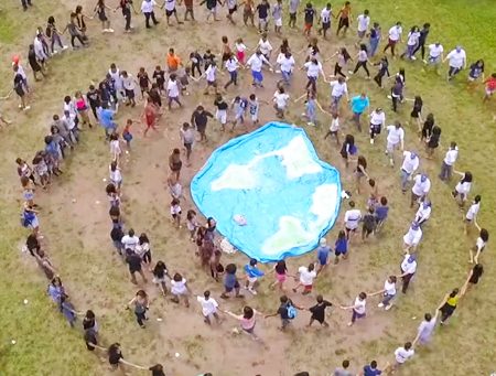 Imagem mostra pessoas em roda. Ao centro algumas pessoas seguram um tecido azul com o mapa múndi