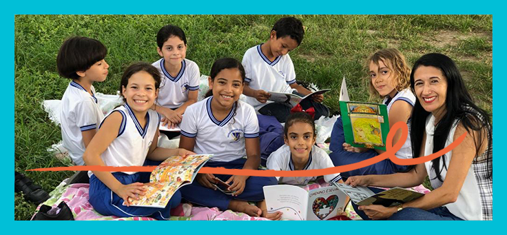 Crianças estão agrupadas em meio a gramado segurando livros e sorrindo, ao lado de educadora que conduz o projeto de estímulo à leitura Piquenique Literário.