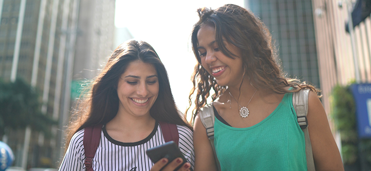 Imagem mostra duas jovens sorrindo enquanto olham para um celular