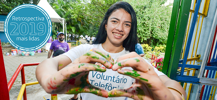 Imagem mostra colaborada do Grupo Telefônica Vivo sorrindo e fazendo um coração com as mãos, que estão sujas de tinta. Ela veste uma camiseta do Programa de Voluntariado da Fundação Telefônica Vivo