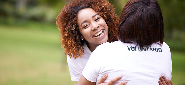 A imagem mostra duas mulheres que participam de programa de voluntariado empresarial estão se abraçando, sendo que é possível ver as costas de uma delas e o rosto sorridente da outra.