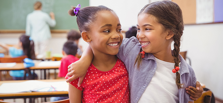 A imagem mostra duas meninas abraçadas e sorrindo uma para outra dentro de uma sala de aula