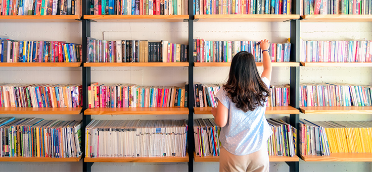 Mulher está de costas para estante com cinco nichos cheios de livros, que ilustram pauta sobre obras essenciais para formação de professores.