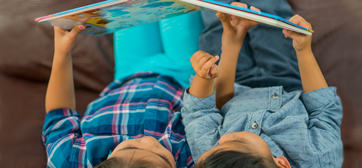 Duas crianças estão lendo deitadas, segurando um livro infantil acima de suas cabeças.