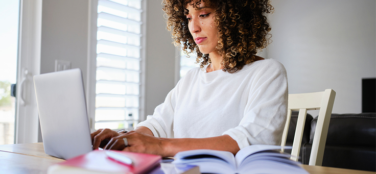 Imagem mostra mulher de blusa branca digitando em um notebook. Na frente dela há um livro aberto.