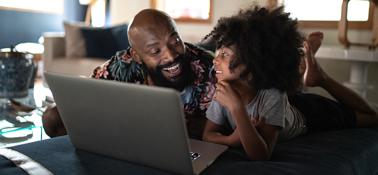 Imagem mostra um pai e uma filha deitados olhando para um notebook e sorrindo