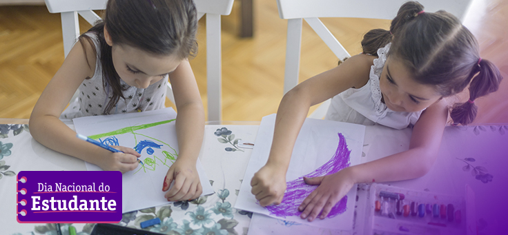 A imagem mostra duas meninas uma ao lado da outra, sentadas atrás de uma mesa. Em cima do móvel, há materiais de desenho e pintura como lápis e giz de cera, e cada uma delas está desenhando em uma folha de papel sulfite (atividades sobre sentimentos)