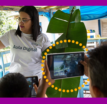 A foto mostra uma formadora do Projeto Aula Digital, que usa uma camiseta com o nome do programa, mostrando uma planta que está em suas mãos para um grupo de alunos. Um deles, segura um tablet com a câmera apontada na direção dela.