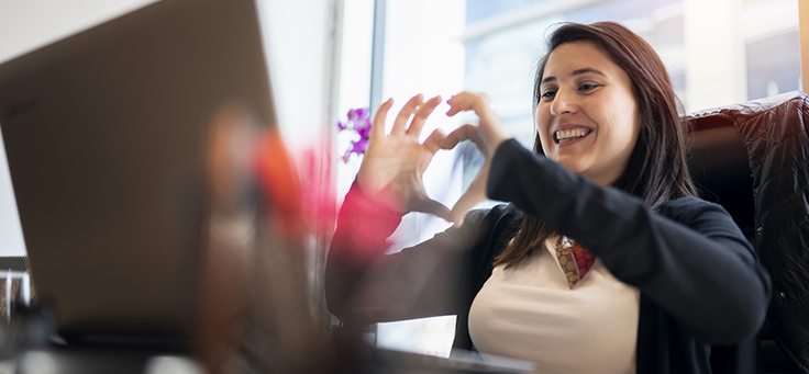 Mulher está sorrindo e olhando para tela do computador enquanto faz sinal de coração com as duas mãos. Imagem ilustra pauta sobre como manter o engajamento e a solidariedade em programas de voluntariado corporativo.