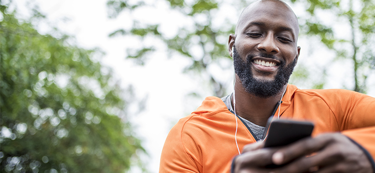 A imagem mostra um homem negro sorrindo, de blusa laranja, usando fones de ouvido enquanto olha para a tela de um celular. Ao fundo dele se vê algumas árvores.