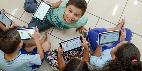 Crianças atendidas pelo projeto Aula Digital ProFuturo em Viamão estão sentadas no chão manuseando os tablets do projeto. Um dos menino está olhando para cima e sorrindo.