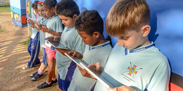 Seis alunos atendidos pelo projeto Aula Digital ProFuturo em Viamão utlizam os tablets do projeto. Eles estão de pé, encostados em um muro da escola.