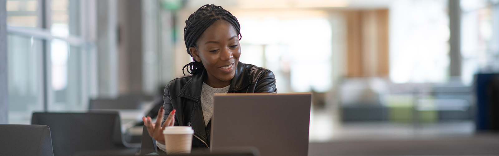 Imagem mostra uma jovem sentada atrás de uma mesa com um notebook a frente dela e um copo de café descartável ao lado