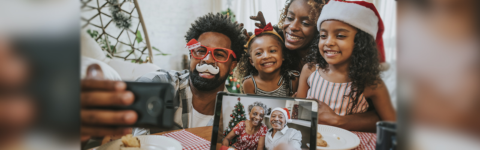 Imagem mostra pai, mãe e filhas fazendo chamada com avós durante o Natal. Os avós aparecem em uma tela de tablet e todos os usam adereços natalinos, como gorro de Papai Noel.