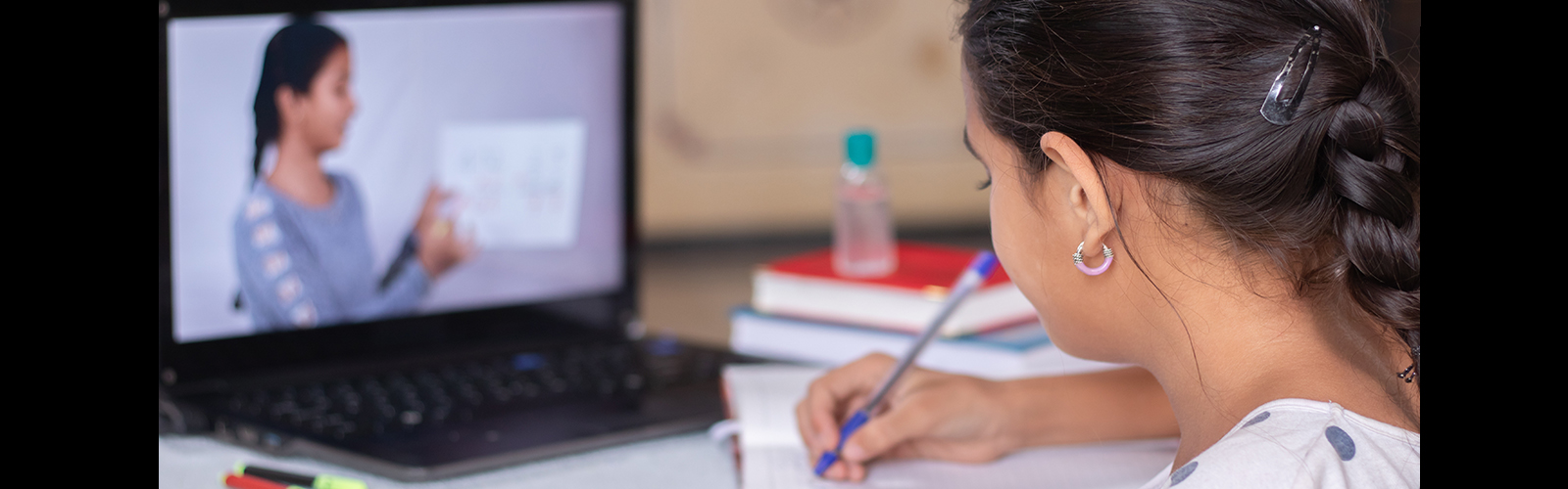 Imagem mostra uma menina sentanda de costas escrevendo em um caderno. Sobre a mesa, há alguns livros e um notebook com a imagem na tela de uma mulher segurando um papel branco.