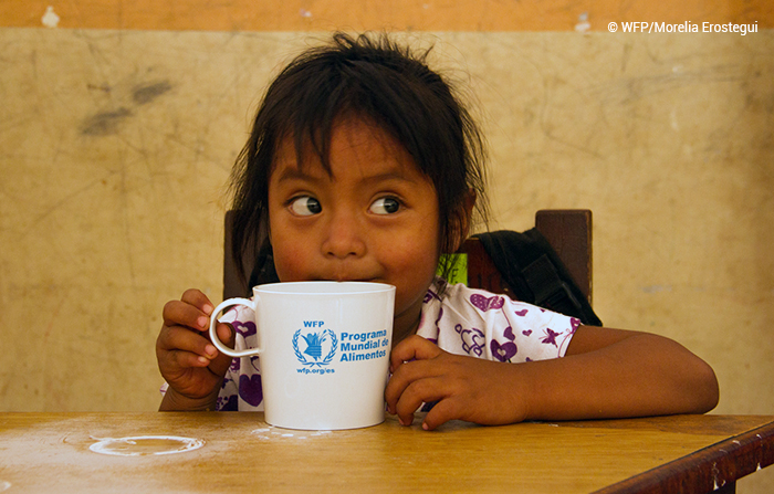 Imagem mostra menina sentada à mesa, com uma caneca onde se lê “Programa Mundial de Alimentos” em azul.