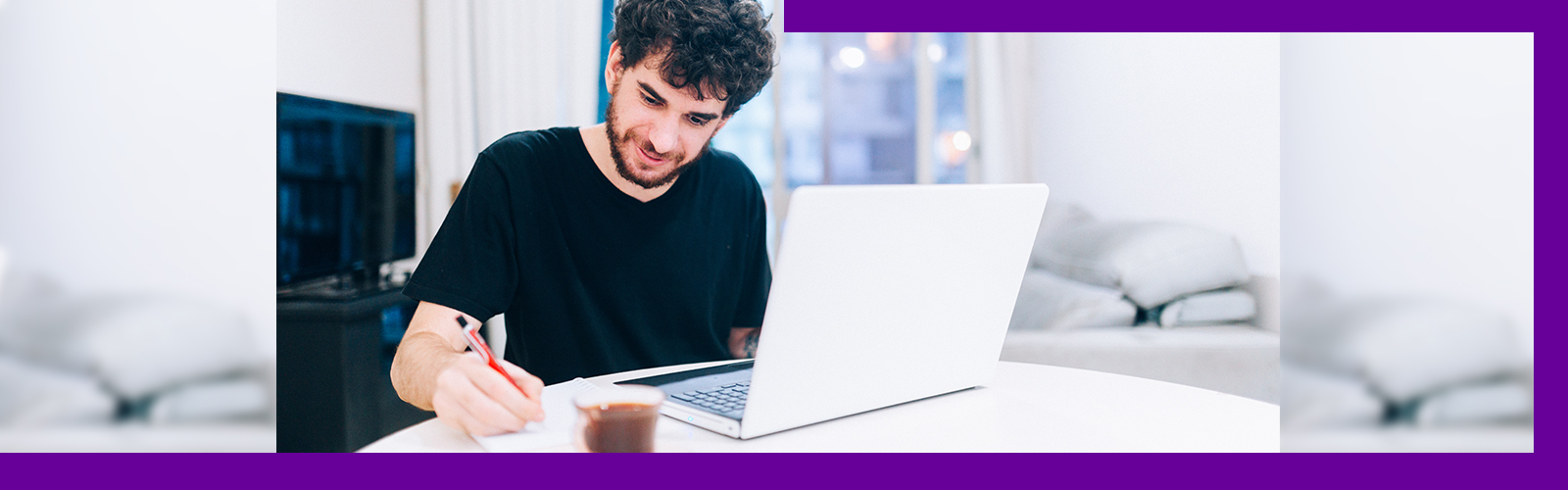 Imagem mostra um jovem de camiseta preta sentado atrás de uma mesa com um notebook a sua frente, fazendo anotações em uma folha.