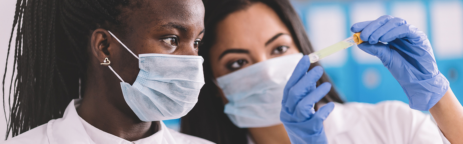 A imagem mostra duas jovens, uma negra e uma branca em um laboratório. Ambas estão com jaleco branco e máscara de proteção e uma delas segura um tubo de ensaio.