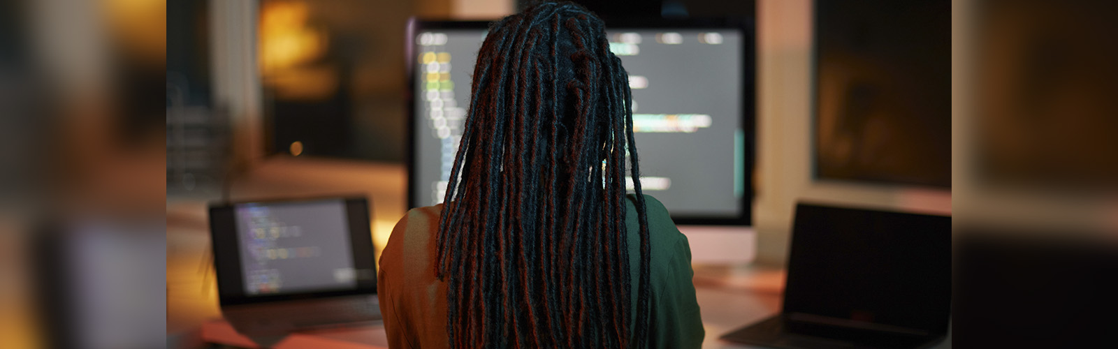 Mulher de dreads trabalha de costas. Na frente dela se vê um notebook e um computador.