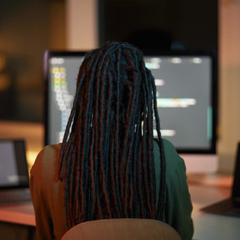 Mulher de dreads trabalha de costas. Na frente dela se vê um notebook e um computador.