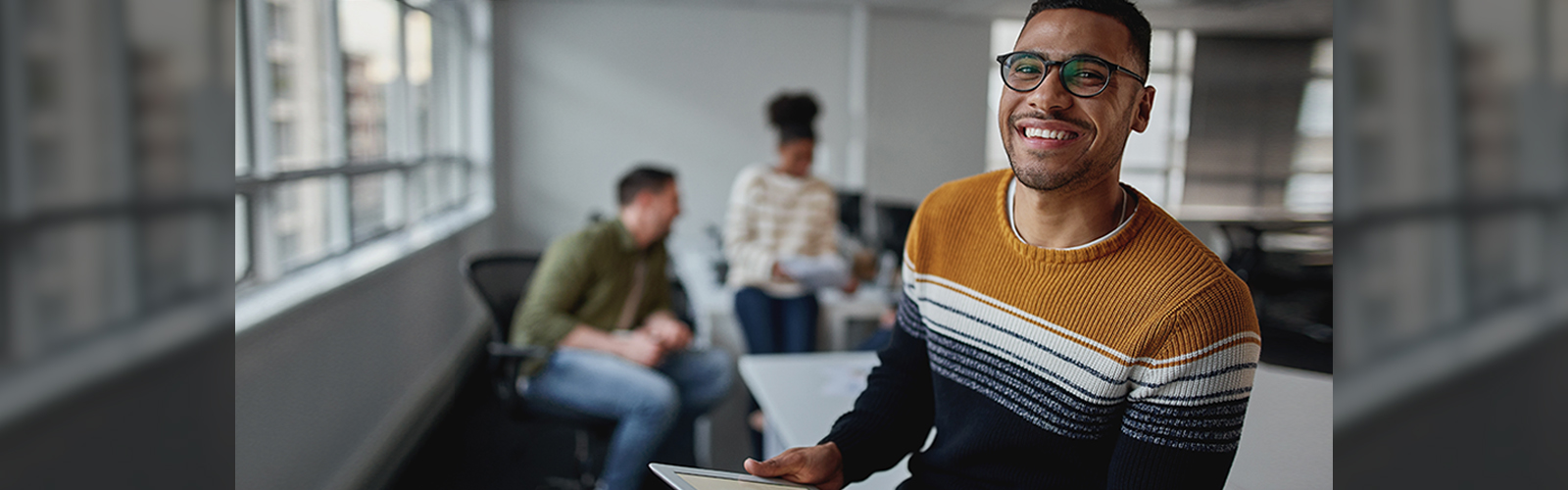 A imagem ilustra pauta sobre transição de carreira e mostra um ambiente de trabalho mais informal. Em primeiro plano há um homem jovem segurando um tablet e sorrindo para a foto. Ele é negro, usa óculos e barba, e está vestindo um suéter com listras nas cores ocre, branco, cinza e preto.