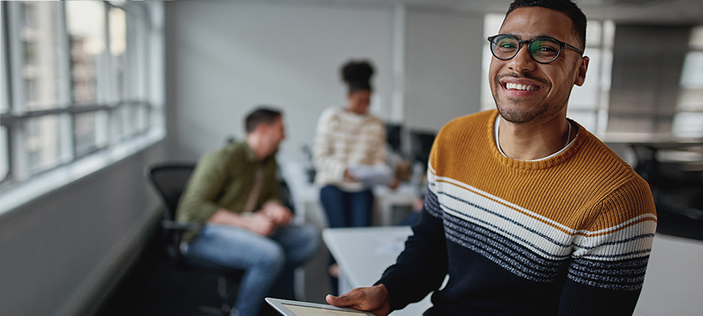 A imagem ilustra pauta sobre transição de carreira e mostra um ambiente de trabalho mais informal. Em primeiro plano há um homem jovem segurando um tablet e sorrindo para a foto. Ele é negro, usa óculos e barba, e está vestindo um suéter com listras nas cores ocre, branco, cinza e preto.