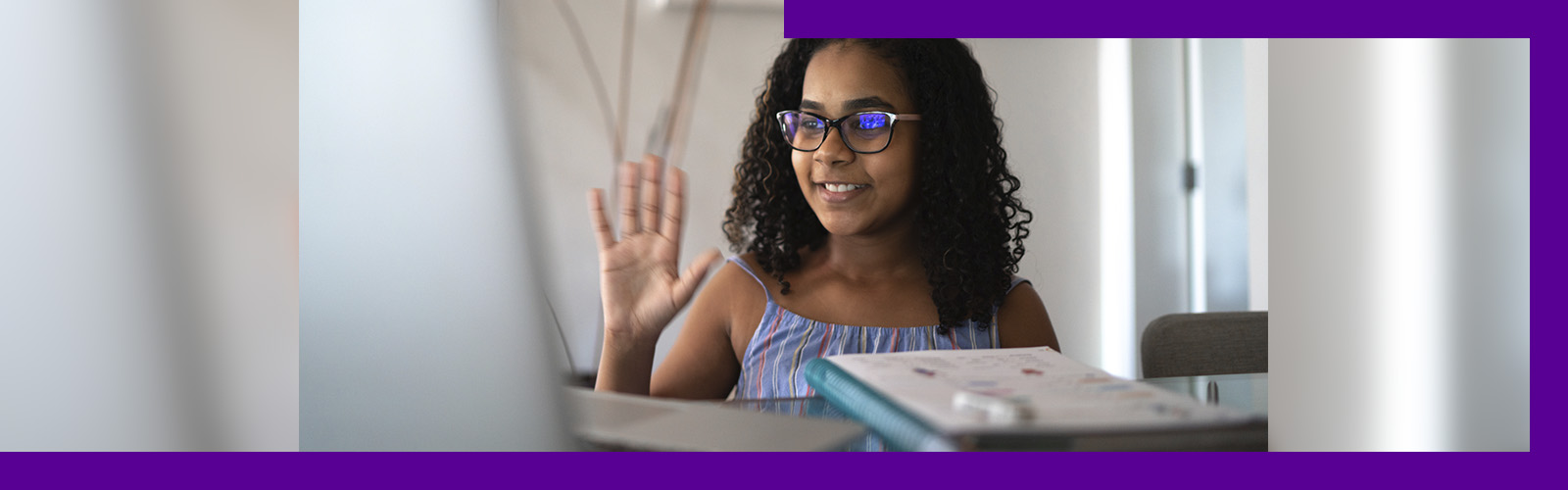 Imagem mostra uma menina sentada em frente a uma mesa onde está um notebook. Ela está sorrindo com uma das mãos levantadas.