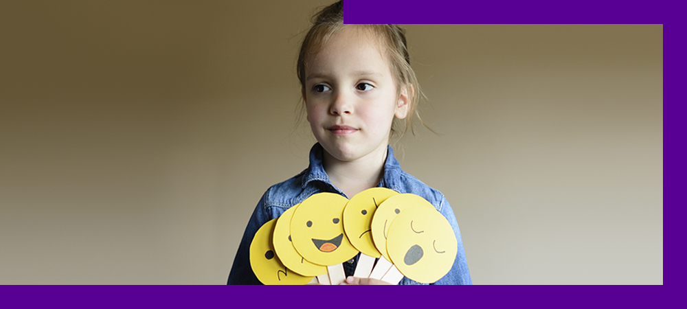 Em imagem que ilustra como ajudar as crianças a se expressarem, uma menina segura diversas placas que lembram emojis de expressões faciais, indicando sono, alegria, tristeza, entre outros. Ela está olhando para a frente com um sorriso discreto.