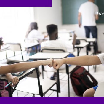 A imagem mostra duas crianças dentro de uma sala de aula, usando máscaras de proteção, se cumprimentando com as palmas das mãos fechadas.