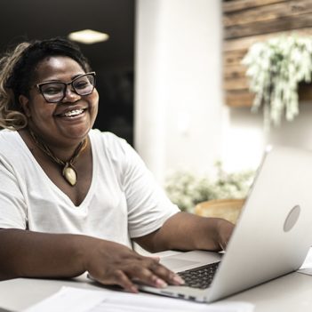 Imagem mostra uma mulher negra, de camiseta branca e um colar. Ela está sentada em frente um computador, sorrindo enquanto olha para a tela.