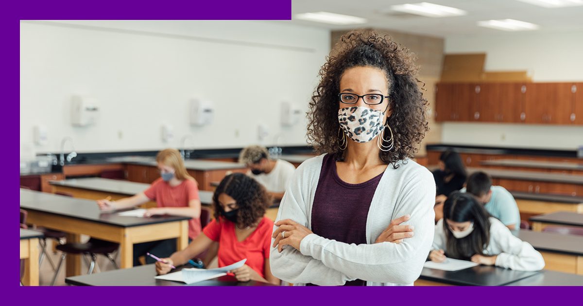 Imagem de uma professora de pé, com os braços cruzados, em uma sala de aula com alunos ao fundo, sentados em carteiras escolares. Todos estão usando máscara de proteção.