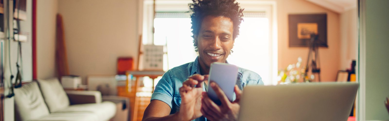 Imagem de um jovem na sala de sua casa, sorrindo enquanto mexe no celular