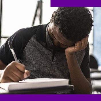 imagem de um jovem estudante na sala de aula, sentando em uma carteira escolar. Ele usa uma máscara de proteção individual e está com uma das mãos apoiada na cabeça, enquanto escreve em um caderno.