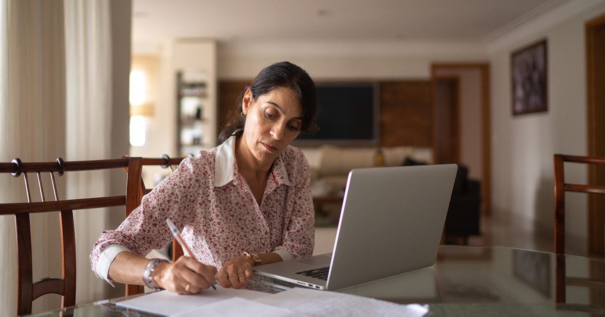 Imagem mostra professora estudando utilizando um notebook e escrevendo em um papel