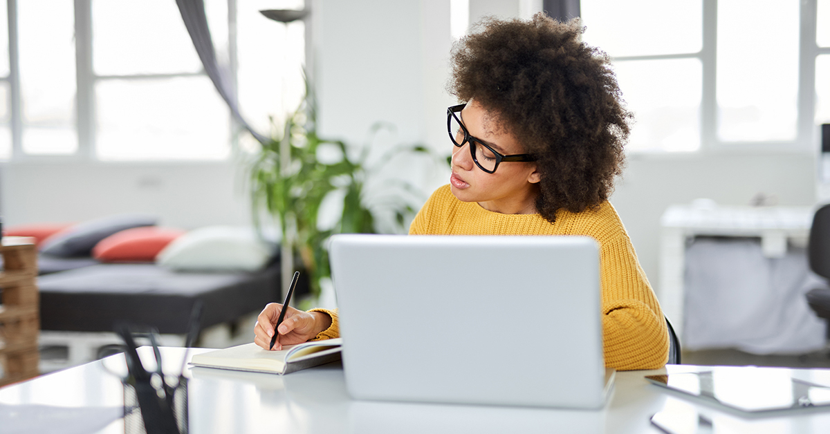 Imagem mostra mulher de óculos estudando em frente a um notebook e usando um caderno e caneta