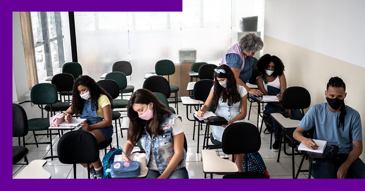 Imagem mostra estudantes em sala de aula sendo orientados por uma professora