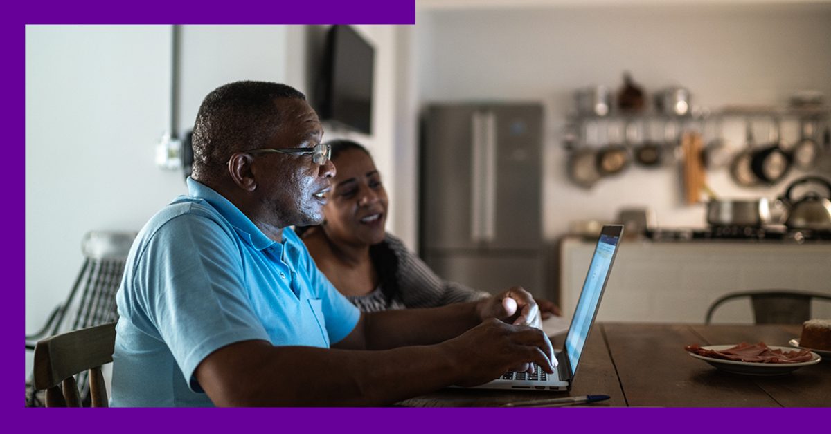 Imagem mostra um homem e uma mulher negros utilizando um computador. Eles estão em casa. O homem veste uma camisa azul. A mulher usa uma blusa cinza e tem cabelos compridos.