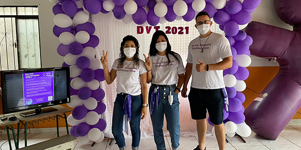 Imagem mostra três voluntários do DVT. Duas mulheres e um homem. Eles estão em frente a um arco de balões nas cores branca e roxa.