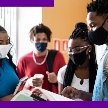 Imagem mostra quatro estudantes negros. Eles estão reunidos e observando o conteúdo de um livro, que está sendo mostrado por um dos garotos. Na imagem há dois garotos e uma garota.