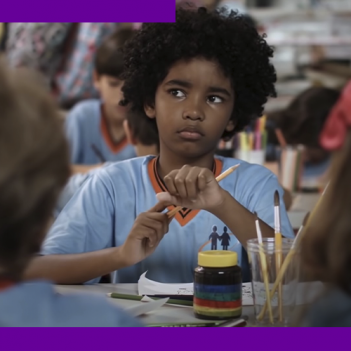 Imagem mostra um menino negro, com uniforme escolar, sentado em uma mesa com outras crianças. Ele segura um lápis de cor. Sobre a mesa, há outros lápis, pincéis, papel e um frasco de tinta