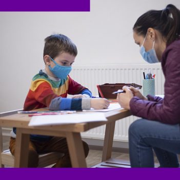 Imagem mostra uma psicopedagoga auxiliando uma criança em sala de aula. A criança está sentado em uma cadeira e em uma mesa há objetos pedagógicos. A profissional está em sua frente.