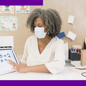 Imagem mostra uma mulher utilizando um tablet. Ela é negra, usa camisa branca e máscara, e está em uma sala com objetos pedagógicos. Ela está sentada e manuseia o tablet em uma mesa