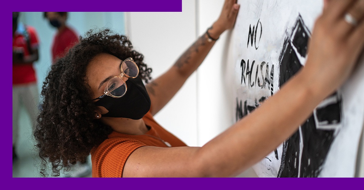 Imagem mostra uma jovem negra fixando em uma parede um cartaz contra o racismo