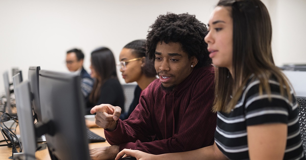 Imagem mostra cinco estudantes em uma sala com computadores. Em primeiro plano, é possível ver um estudante e uma estudante conversando enquanto olham para a tela do computador. O jovem é negro e usa uma blusa na cor vinho e a jovem é branca e uma camiseta listrada.