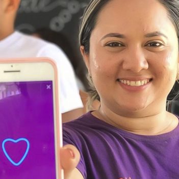 Imagem mostra a voluntária Geciclelma. Ela usa uma camiseta na cor roxa, está segurando um celular com a mão direita e mostrando ele para a câmera. Ela está sorrindo. Na tela do celular, há uma imagem na cor roxa com a ilustração de um coração.