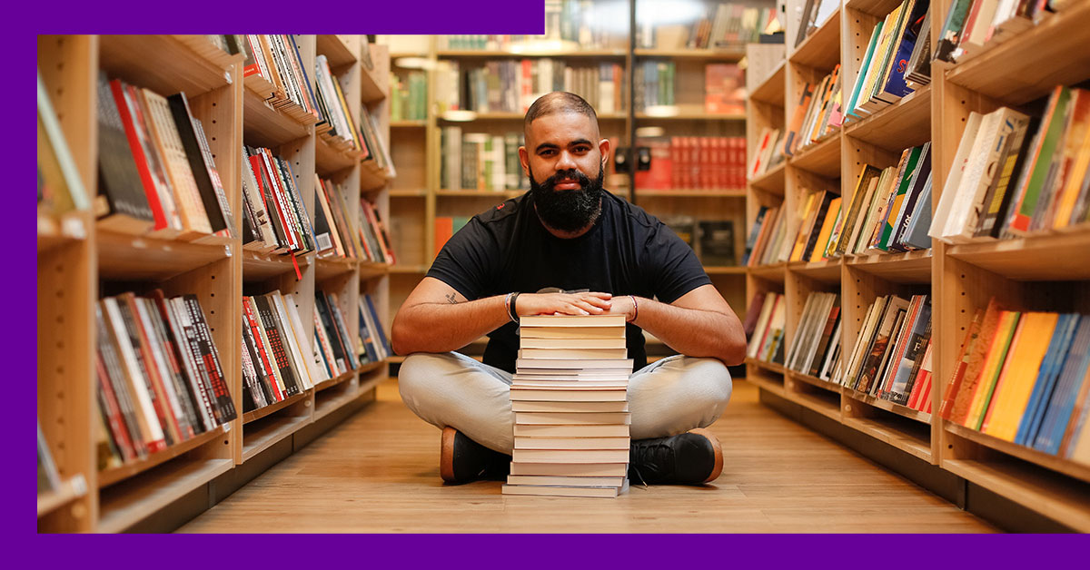 O empreendedor social Tiago Silva está sentando no chão de uma biblioteca, com as pernas cruzadas. A sua frente uma pilha de livros, na qual ele está com as mãos apoiadas.